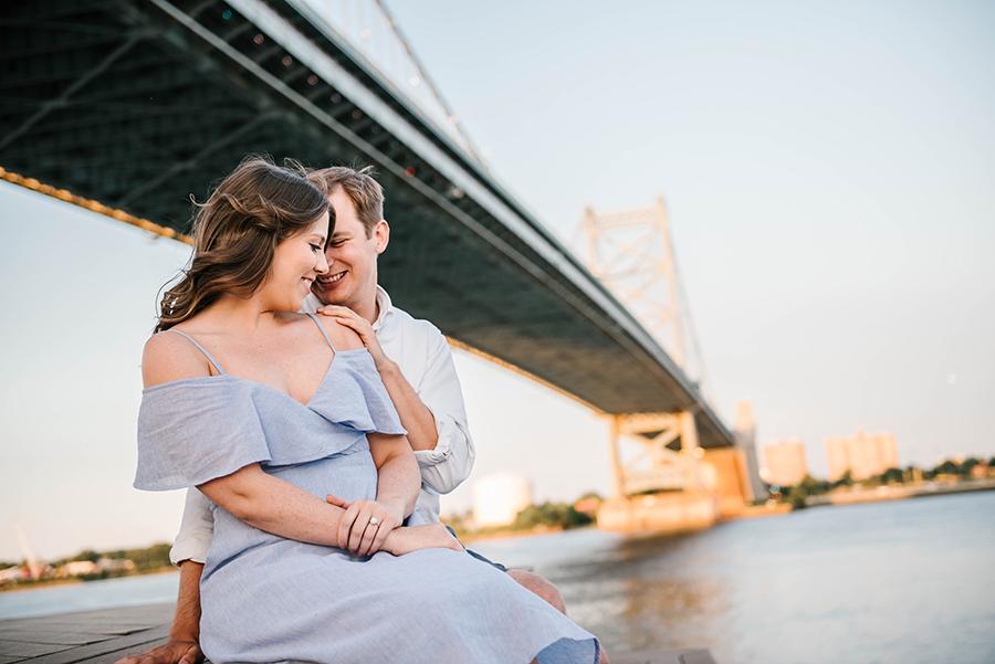 couple embrace in front of Ben Franklin Bridge with engagement ring by Nicole Cordisco and Philly In Love