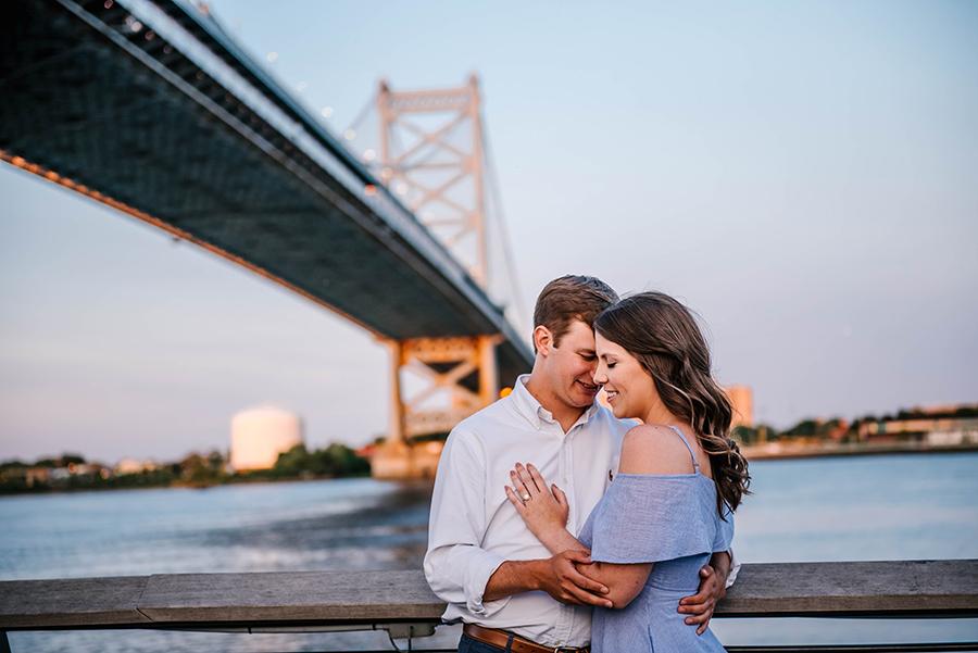 couple embrace in front of Ben Franklin Bridge with engagement ring by Nicole Cordisco and Philly In Love